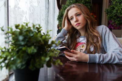 En flicka som tittar ut genom ett fönster. Hon har brunt långt hår och en grå tröja på sig. I den vänstra handen håller hon i en smartphone.