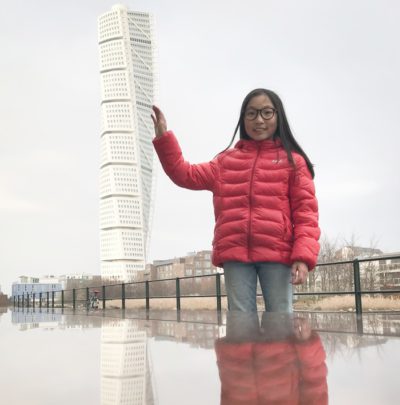 Alma håller sin hand mot byggnaden Turning torso. Alma är nära kameran, byggnaden är långt borta, ändå ser det ut som att Alma lutar handen mot höghuset.