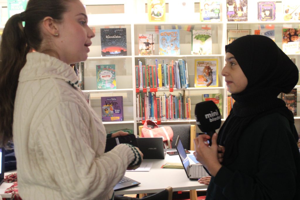 En elev på reporterskolan intervjuar koncentrerat sin lärare och håller i en mick som det står Minibladet på.